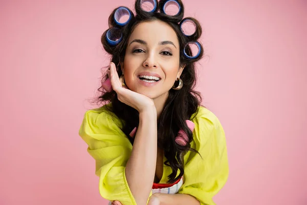 stock image portrait of brunette woman in yellow blouse and hair curlers holding hand near face and looking at camera isolated on pink