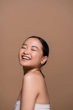 Joyful asian woman in top closing eyes while posing isolated on brown 