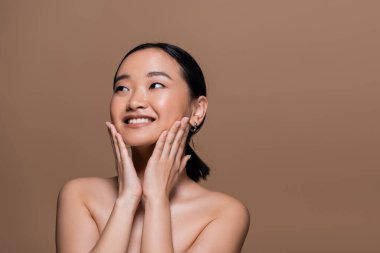 Cheerful asian model with naked shoulders touching cheeks isolated on brown 