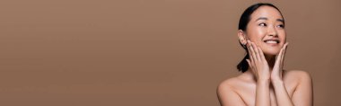 Positive asian woman with makeup and naked shoulders touching cheeks isolated on brown, banner 
