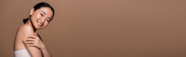 Cheerful asian woman in top touching arm and looking at camera isolated on brown, banner 