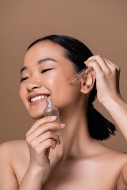 Positive asian model with naked shoulders applying serum on face isolated on brown 