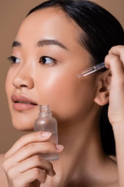 Young asian woman applying serum on cheek isolated on brown 