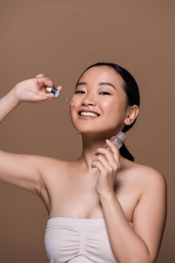 Cheerful asian woman holding bottle and pipette of cosmetic serum isolated on brown 