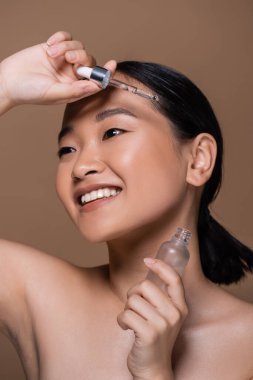 Smiling asian woman with naked shoulders holding cosmetic serum isolated on brown 