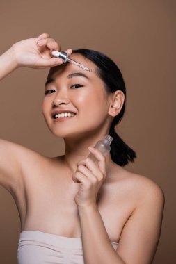 Pleased asian woman in top holding pipette and bottle of cosmetic serum isolated on brown 