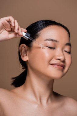 Joyful asian woman with dropper applying cosmetic serum on cheek isolated on brown 