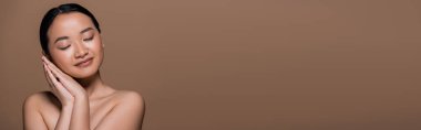 Pretty asian woman with naked shoulders holding hands near cheek isolated on brown, banner 