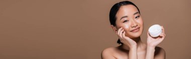 Smiling asian model applying face cream on cheek isolated on brown, banner 