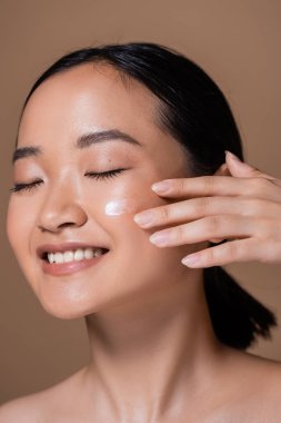 Smiling asian woman applying face cream isolated on brown 