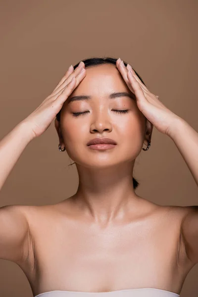 Young asian woman in top touching forehead isolated on brown