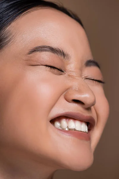Close up view of happy asian woman with natural makeup closing eyes isolated on brown