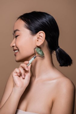 Side view of smiling asian woman with naked shoulders holding jade roller isolated on brown 