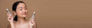 Brunette asian woman with naked shoulders holding bottles of cosmetic serums isolated on brown, banner 