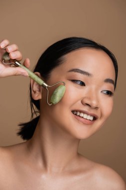 Positive asian model with naked shoulders using jade roller on face isolated on brown 