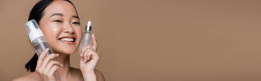 Cheerful asian woman holding face cleanser and serum isolated on brown, banner 