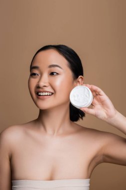 Cheerful asian model in top holding cosmetic cream isolated on brown 