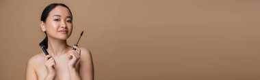 Young asian woman with naked shoulders holding mascara isolated on brown, banner 