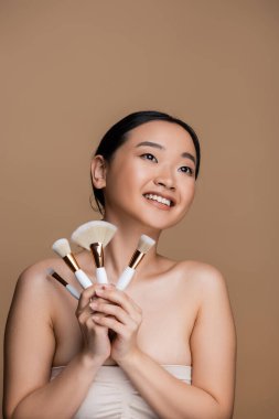 Positive asian model in top holding cosmetic brushes isolated on brown 