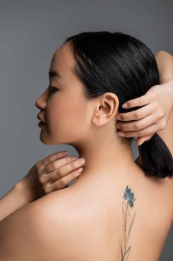 Side view of tattooed asian woman touching hair isolated on grey 