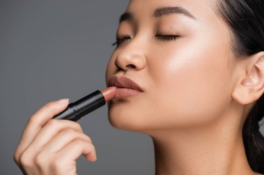 Young asian woman with makeup applying lipstick isolated on grey 