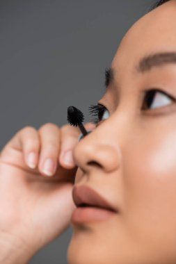 close up view of young asian woman applying black mascara isolated on grey clipart
