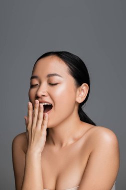 young asian woman with perfect skin and bare shoulders covering mouth with hand while yawning with closed eyes isolated on grey clipart