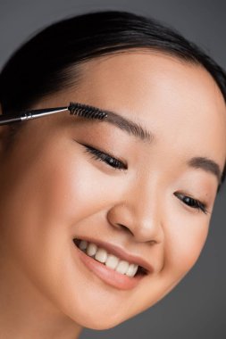 portrait of cheerful asian woman with natural makeup brushing eyebrow isolated on grey