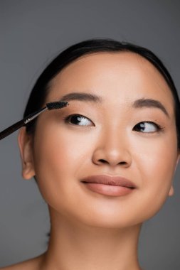 portrait of young asian woman with perfect skin brushing eyebrow isolated on grey