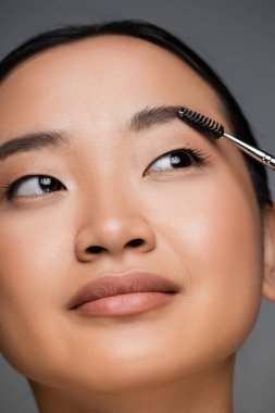 close up portrait of pretty asian woman brushing eyebrow isolated on grey
