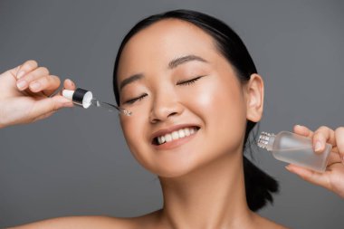 cheerful asian woman with closed eyes and perfect skin holding bottle and pipette of cosmetic serum isolated on grey