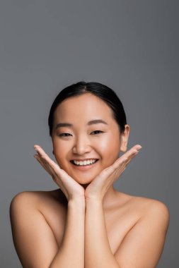 asian woman with natural makeup holding hands near face and looking at camera isolated on grey