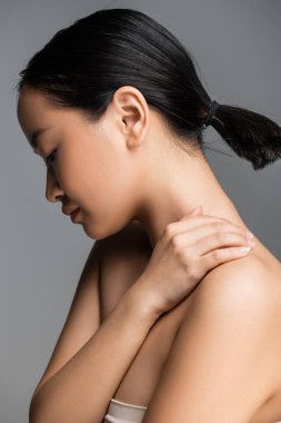 side view of asian woman with ponytail posing with hand on naked shoulder isolated on grey