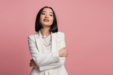 brunette asian woman in white jacket and necklaces standing with crossed arms and looking away isolated on pink