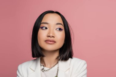 portrait of young asian woman with blue eyeliner and silver necklaces looking away isolated on pink