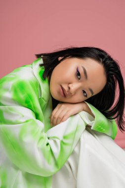 portrait of young asian woman in green and white outfit and visage with blue eyeliner looking at camera isolated on pink