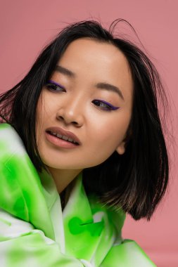 portrait of brunette asian woman with blue eyeliner looking away isolated on pink