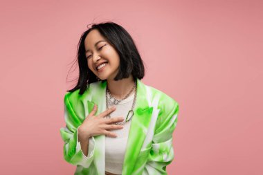 excited and trendy asian woman holding hand on chest while laughing with closed eyes isolated on pink clipart