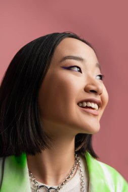 portrait of cheerful asian woman in metal necklaces and makeup looking away isolated on pink