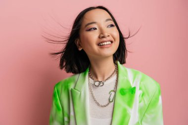 joyful asian woman in trendy outfit and makeup with blue eyeliner looking away on pink background clipart
