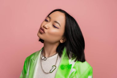 flirty asian woman in metal necklaces and trendy jacket pouting lips isolated on pink