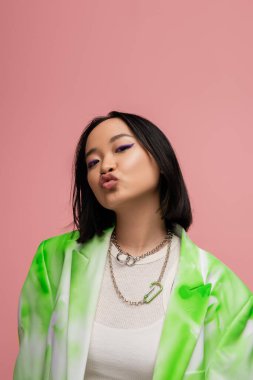 flirty and trendy asian woman in metal necklaces and blazer pouting lips while looking at camera isolated on pink