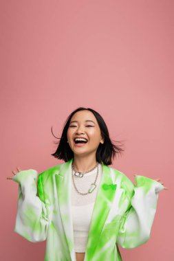 cheerful and stylish asian woman in fashionable jacket and necklaces showing wow gesture isolated on pink