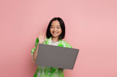 joyful and trendy asian woman waving hand during video call on laptop isolated on pink