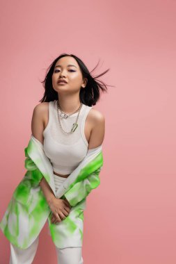 brunette asian woman in white tank top and stylish jacket looking at camera isolated on pink