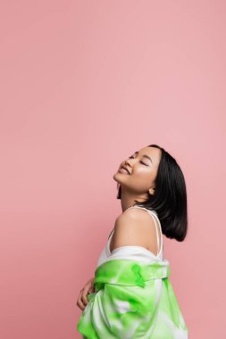 pleased asian woman with closed eyes posing with green and white jacket isolated on pink
