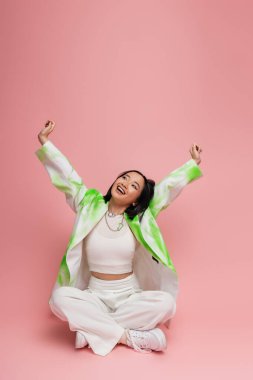 joyful asian woman in stylish outfit sitting with crossed legs and stretching raised hands on pink background