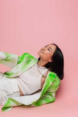 happy asian woman in fashionable outfit and necklaces looking away while lying on pink background