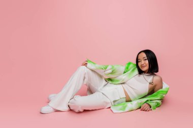 full length of smiling asian woman in white pants and fashionable jacket lying on pink background