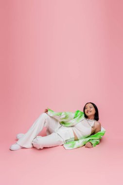 full length of asian model in white pants and trendy jacket lying and looking at camera on pink background